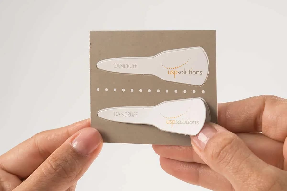 Hands Holding Card with Two White Dandruff Test Strips Labeled 'uspsolutions' on Gray Background