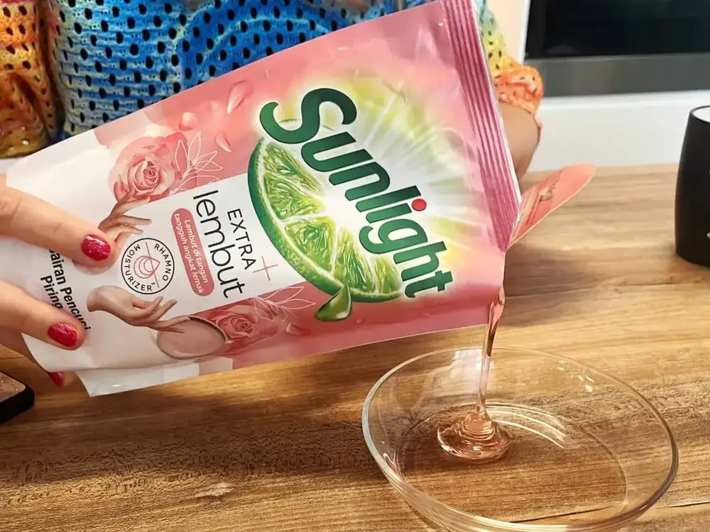 Pouring Pink Liquid Dish Soap from Sunlight Packaging into Clear Glass Bowl on Wooden Table with Colorful Knit Sweater