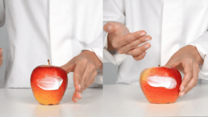 A person wearing a white coat applies and spreads white cream on a red apple, showing the apple before and after