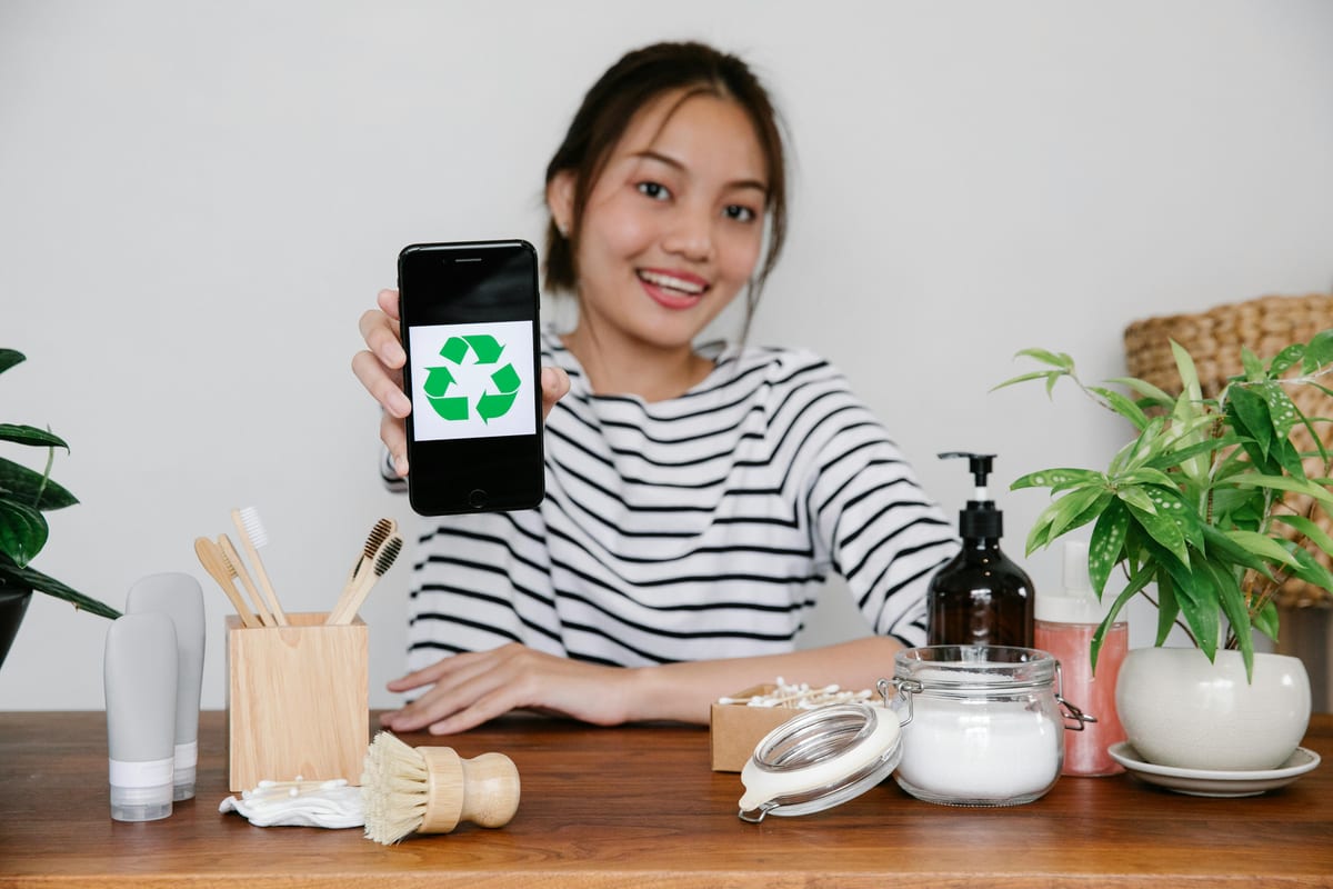 Smiling Asian woman demonstrating recycling symbol on smartphone while sitting at table with eco friendly cosmetic