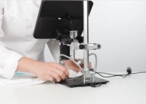 A person in a white lab coat adjusts a digital microscope with a screen on a white table, preparing for a scientific or