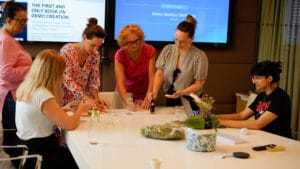 Five women collaborate at a conference table with papers, pens, and glasses. Two large screens display workshop