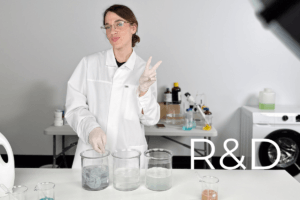 A woman in a lab coat and gloves stands behind three large beakers with clear liquids, making a peace sign. Lab