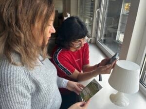 Two women stand by a window; one holds a phone and scans a code on a lamp, while the other holds a pamphlet with green