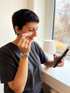 A person with short dark hair smiles while applying a cream to their cheek, holding a phone in their other hand. They